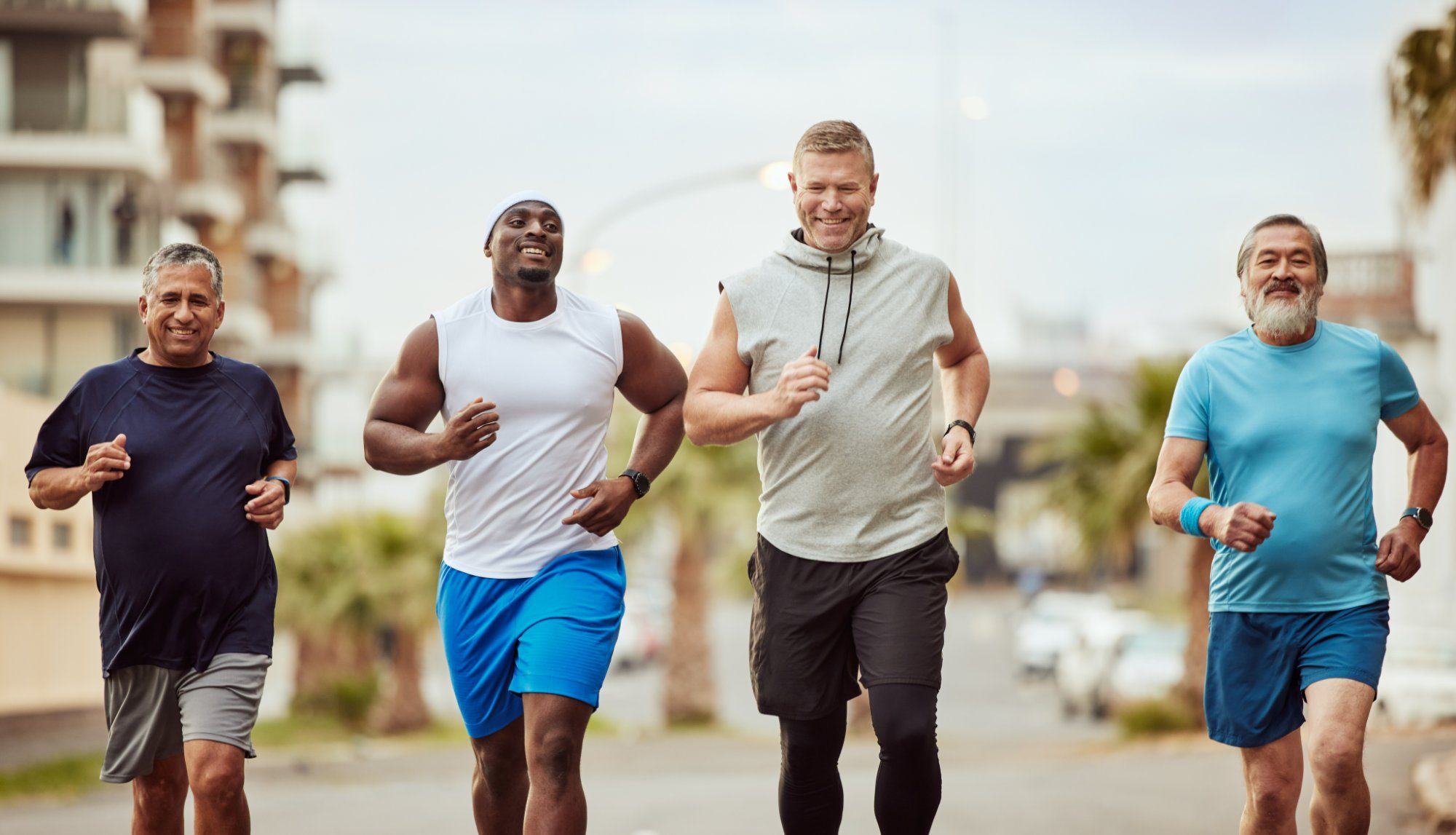Four men in their 40s to 60s jogging together down a city street, smiling